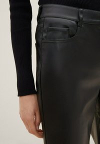 Vue de profil d'une personne portant un pantalon noir en similicuir et un haut noir côtelé à manches longues, avec la main visible détendue le long du corps.