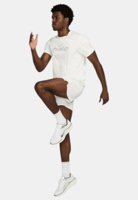 Camiseta blanca de Nike con un logo gris, combinada con pantalones cortos blancos y zapatillas Nike. El sujeto está en una pose de carrera dinámica, mostrando un movimiento atlético.