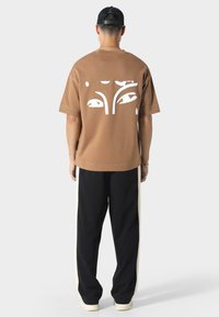 Brun oversized t-shirt med et hvidt abstrakt design på ryggen, parret med sorte bukser med hvide sideløsninger.