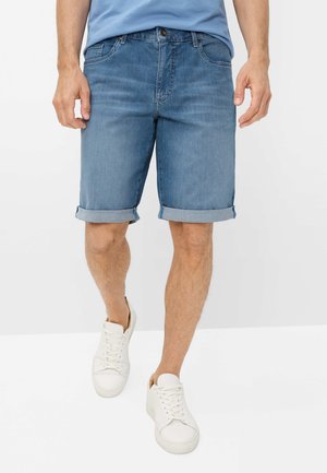 Mann trägt hellblaue hochgekrempelte Jeansshorts, weiße Sneaker und ein hellblaues Hemd und steht vor einem schlichten weißen Hintergrund.