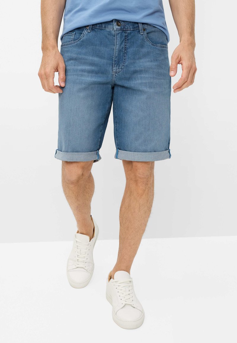 Homme portant un short en jean bleu clair retroussé, des baskets blanches et une chemise bleu clair, debout devant un fond blanc uni.
