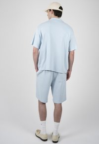 Homme portant une chemise bleu clair à manches courtes et un short, casquette de baseball beige, chaussettes blanches et mocassins beiges, debout de dos sur un fond uni.