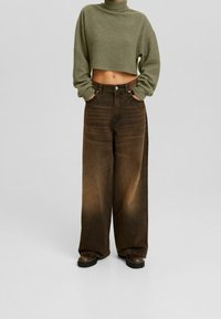Zkrácený olivově zelený svetr s vysokým límcem, doplněný o široké hnědé džínové kalhoty s vybledlou texturou a detaily kapes.