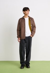 Bruine gevoerde jas met een kraagontwerp over een wit T-shirt, gecombineerd met losse zwarte jeans en zwarte schoenen, op een groene vloer.