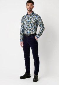Bloemen langemouw shirt in blauw en groen op een witte achtergrond, gecombineerd met donker marineblauwe corduroy broek en zwarte enkellaarsjes.