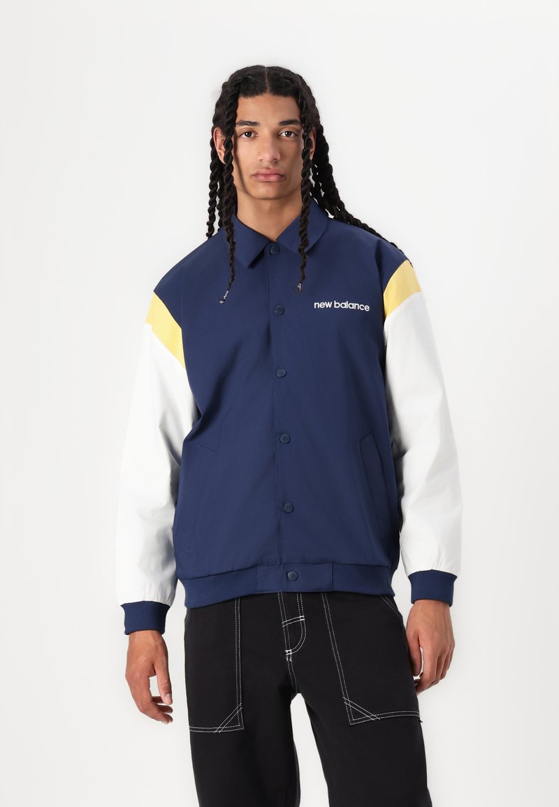 Veste bleu marine avec des manches blanches et jaunes ; comporte des boutons, un col et des poches latérales ; fabriquée dans un tissu lisse et texturé.