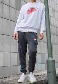Vitt sweatshirt med rosa Nike-logotyp, grå cargobyxor med logotypdetaljer och ljusgrå sneakers med perforerade detaljer, stående på en stenyta.