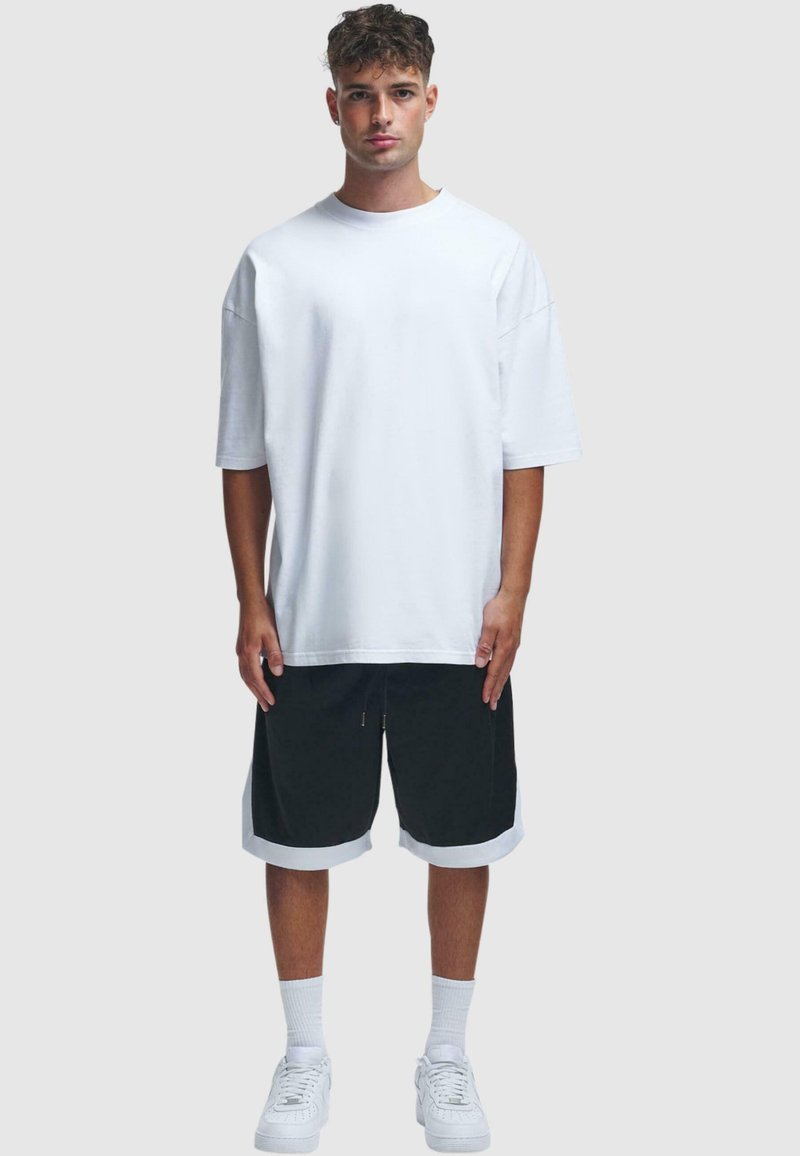 Fehér, oversize T-shirt rövid ujjal, fekete sorttal, fehér díszítéssel, és fehér sportcipővel párosítva. Sima textúra, laza szabás.