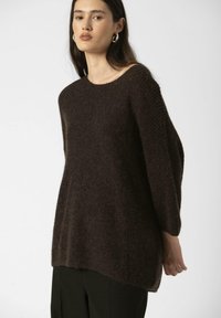 Pull en tricot marron surdimensionné avec une texture douce et un col rond, présentant des épaules tombantes et des fentes sur les côtés. Porté sur un pantalon noir.
