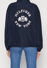 Granatowa kurtka wiatrówka o luźnym kroju, z dużym białym logo "Hilfiger New York" na plecach i elastycznymi mankietami dla wygody.