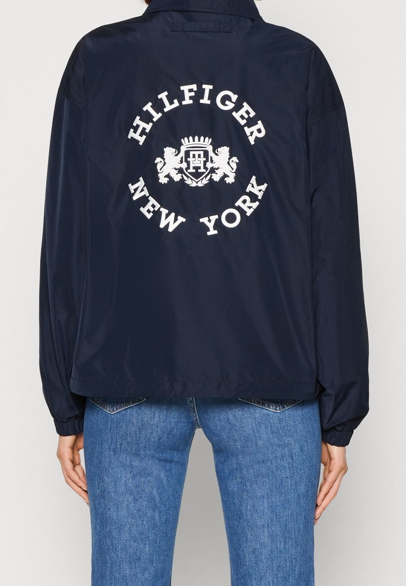 Granatowa kurtka wiatrówka o luźnym kroju, z dużym białym logo "Hilfiger New York" na plecach i elastycznymi mankietami dla wygody.