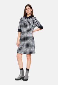 Kurzes Kleid mit Kragen, navy-weißem geometrischem Muster, zwei Fronttaschen mit kontrastierendem Besatz, getragen mit grauen Stiefeletten.