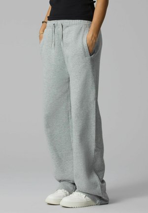 Personne portant un pantalon de jogging ample gris clair avec un cordon et des baskets blanches, mains dans les poches, sur un fond gris uni.