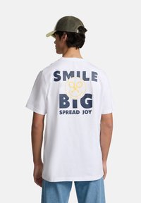 Camiseta blanca con un gran texto en la parte de atrás: "SONRÍE GRANDE PROPAGA ALEGRÍA" con una carita sonriente amarilla y letras en azul oscuro. Corte casual.