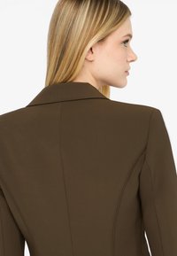 Blazer marrone con colletto strutturato, texture liscia e cuciture sartoriali, caratterizzato da un design aderente con dettagli sul retro e senza hardware visibile.