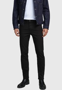 Jeans negros con un diseño ajustado, hechos de mezclilla. Presentan una bragueta con botones, cinco bolsillos y una textura suave. Llevados con una camisa blanca y una chaqueta de mezclilla.