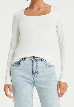 Femme portant un haut blanc à manches longues avec un col carré et un jean taille haute bleu clair, visible du cou aux hanches.