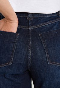Jeans de mezclilla azul oscuro con forma ajustada, costuras visibles y dos bolsillos traseros. La textura de la tela parece suave y resistente.