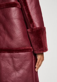 Bordeaux frakke med en tekstureret læderoverflade og bløde, luksuriøse manchetter, synlige sømme og en glat finish.