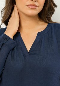 Blusa blu navy con scollatura a V, maniche lunghe e polsini con bottoni. Il tessuto presenta una sottile trama con una leggera lucentezza.