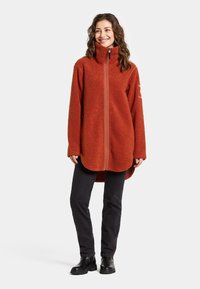 Rustfarvet fleecejakke med høj krave, fuld lynlås og lange ærmer. Har en rund bund og struktureret stof. Sorte bukser og støvler.