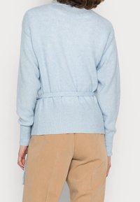 Jasnoniebieski sweter z dzianiny z prążkowanym wykończeniem u dołu i na rękawach, z paskiem w talii i teksturowanym wykończeniem. Umiejscowiony na beżowych spodniach.