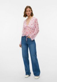 Blouse rose légère à imprimé floral, à manches longues et fermeture à nouer, associée à un jean bleu taille haute et des chaussures blanches.
