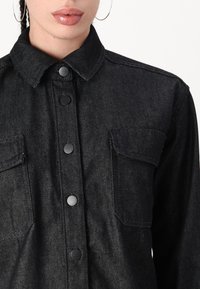 Camisa de mezclilla negra con cuello puntiagudo, cierre frontal de botones, dos bolsillos en el pecho y una tela texturizada sutil. Presenta botones negros.