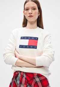 Λευκή μπλούζα με λογότυπο "TOMMY JEANS" σε ναυτικό μπλε, κόκκινο και λευκό διαγωνίως στην κοιλιά. Ριμπ cuffs και στρίφωμα, μαλακό βαμβακερό υλικό. Κόκκινο καρό πουκάμισο δεμένο γύρω από τη μέση.