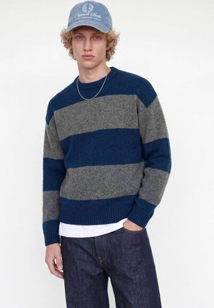 Jeune homme portant un pull rayé bleu et gris, un jean foncé, une casquette bleu clair et un collier en chaîne argentée, debout avec une main dans la poche.