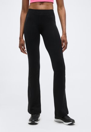 Kvinde iført sorte vide bukser, sorte atletik-sko og en pink cropped top mod en ensartet lys baggrund.