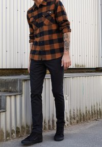 Bruin en zwart geblokt flanellen shirt met twee borstzakken, gecombineerd met zwarte slim-fit broeken en zwarte sneakers. Betonnen trappen op de achtergrond.