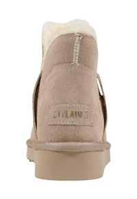 Bota de invierno de ante beige con forro de piel de oveja, costuras visibles y suela plana, mostrada desde la parte trasera con una etiqueta de marca cosida.