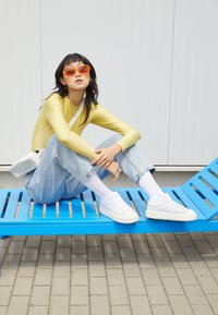 Haut à manches longues jaune, jean baggy bleu clair, baskets blanches et lunettes de soleil orange. Assis sur un banc à lattes bleu contre un mur blanc.