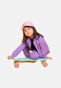 Dziecko w fioletowej kurtce i spodniach klęczy na jasnozielonym deskorolce z różowymi kołami. Ma na sobie różową czapkę i długie ciemne włosy.