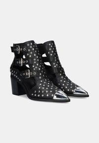 Botas de tobillo de cuero negro con tachuelas plateadas, recortes y dos correas con hebilla. Presentan una puntera puntiaguda con punta metálica y un tacón ancho.