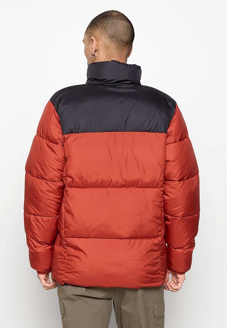 Mężczyzna w czerwonej i czarnej kurtce puchowej widocznej od tyłu, noszący oliwkowe spodnie cargo na jednolitym białym tle.