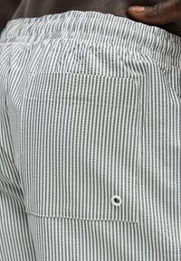 Τσέπη σε ριγέ σορτς με κάθετες μαύρες και λευκές γραμμές, στερεωμένη με ελαστική μέση, με ένα χέρι που στηρίζεται στο ισχίο.