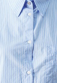Light blue button-up shirt with thin white stripes, featuring a classic collar, a single chest pocket, and white buttons. Soft cotton texture.