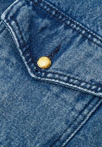 Zbliżenie na tkaninę dżinsową z złotym guzikiem, niebieskimi ściegami i lekko zniszczoną fakturą w okolicy kieszeni.