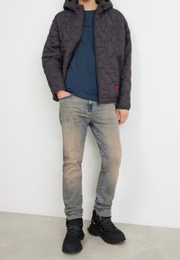 Steppjacke in Dunkelgrau mit Kapuze, Reißverschluss vorne und seitlichen Taschen, kombiniert mit einem blauen T-Shirt und hellblauen Jeans sowie schwarzen Sneakers.