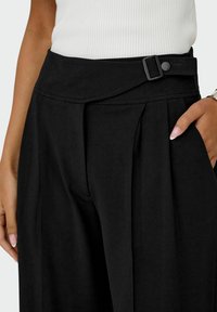 Černé kalhoty s vysokým pasem a přehnutým pasovým lemem, zdobené bočním knoflíkem a záhyby. Hladká textura látky.