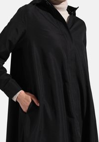 Manteau noir léger avec capuche, doté d'une fermeture éclair frontale, de manches longues et d'un léger éclat. Comprend des poches latérales pour plus de fonctionnalité.