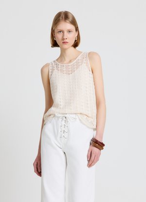 Jeune femme portant un haut en dentelle beige sans manches, un pantalon blanc taille haute avec un laçage à l'avant, et des bracelets marron à son poignet droit.