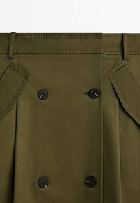 Gros plan sur un tissu vert olive avec quatre boutons noirs et des rabats de poche cousus en biais de chaque côté.