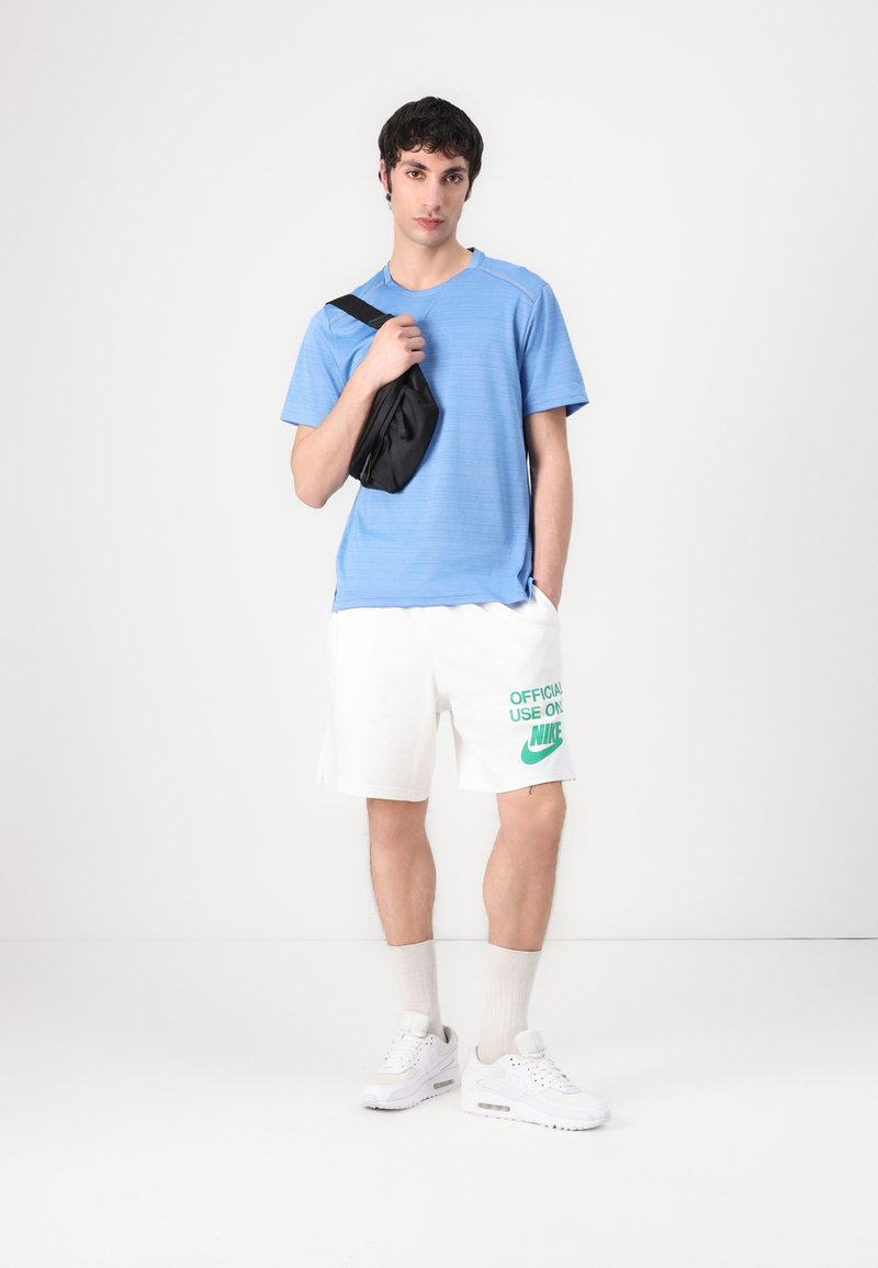 Camisa de manga corta azul claro, pantalones cortos blancos con texto verde "NIKE", y zapatillas blancas. El modelo sostiene un pequeño bolso negro de hombro.