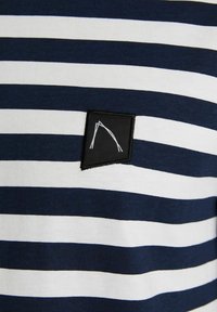 Jūros mėlynumo ir baltų horizontalių juostelių audinys su maža juoda pleistra, ant kurio pritvirtintas baltas minimalistinis arko dizainas.