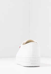 Baskets en toile blanche avec une semelle plate en caoutchouc. Elles présentent une étiquette logo rouge au talon et une tige texturée pour une meilleure adhérence et durabilité.