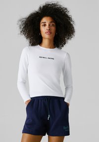 Kobieta o kręconych włosach, ubrana w białą koszulkę z długim rękawem "Red Bull Racing" oraz granatowe krótkie spodenki, z rękami w kieszeniach.