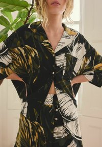 Camisa de botones con patrón en negro y dorado, con mangas anchas, combinada con pantalones cortos a juego. La tela presenta diseños audaces y abstractos de hojas.